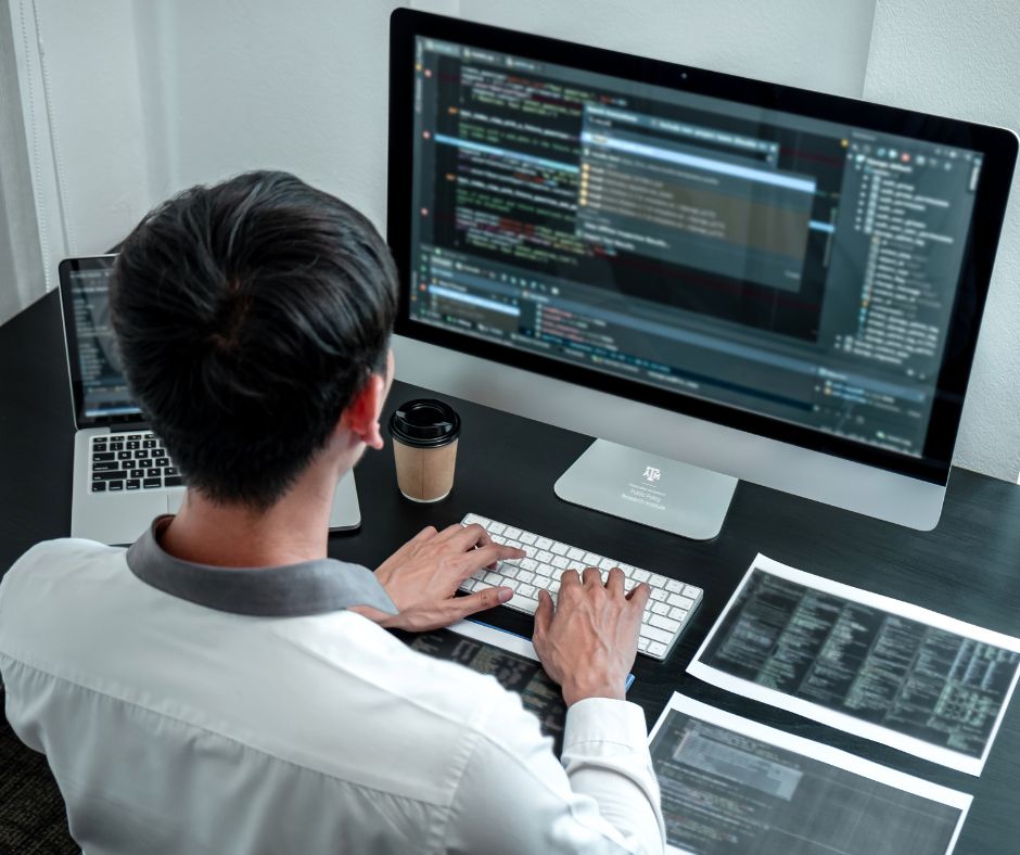 Person working at a desk on a large computer monitor displaying multiple lines of code, with a keyboard, coffee cup, and printed documents nearby.
